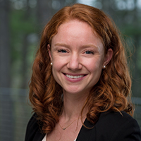Professional headshot of a smiling woman with red hair wearing a black blazer.