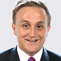 Headshot of a smiling man in a suit and tie against a light gray background.