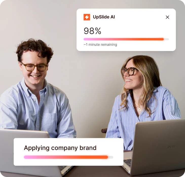 Two colleagues using UpSlide AI to apply company branding on laptops, smiling and collaborating.