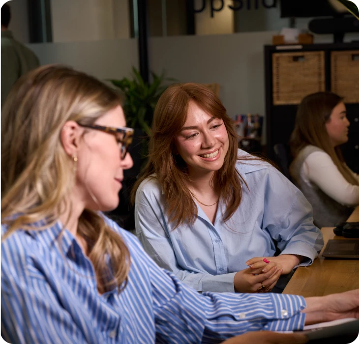 Two women in blue shirts collaborating at an office desk, smiling and discussing work.
