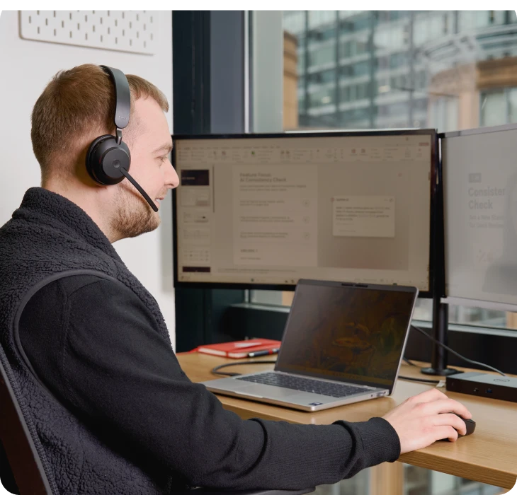 Man wearing headset working at a desk with laptop and dual monitors.