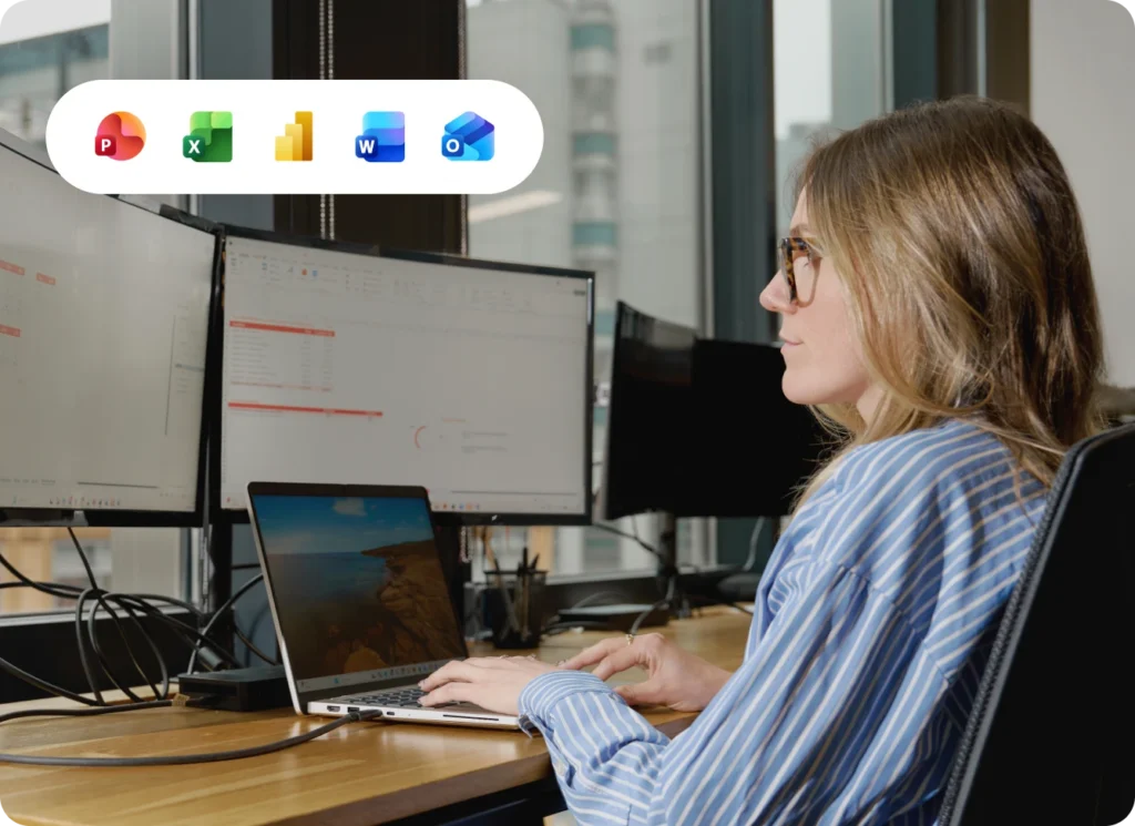 Woman using laptop with Microsoft Office icons. Two monitors on desk in office setting.